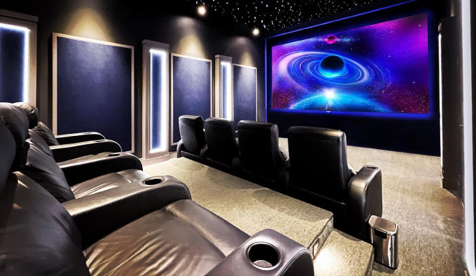 Home theater room with black reclining seats, illuminated wall panels, starry ceiling, and screen showing galaxy with black hole